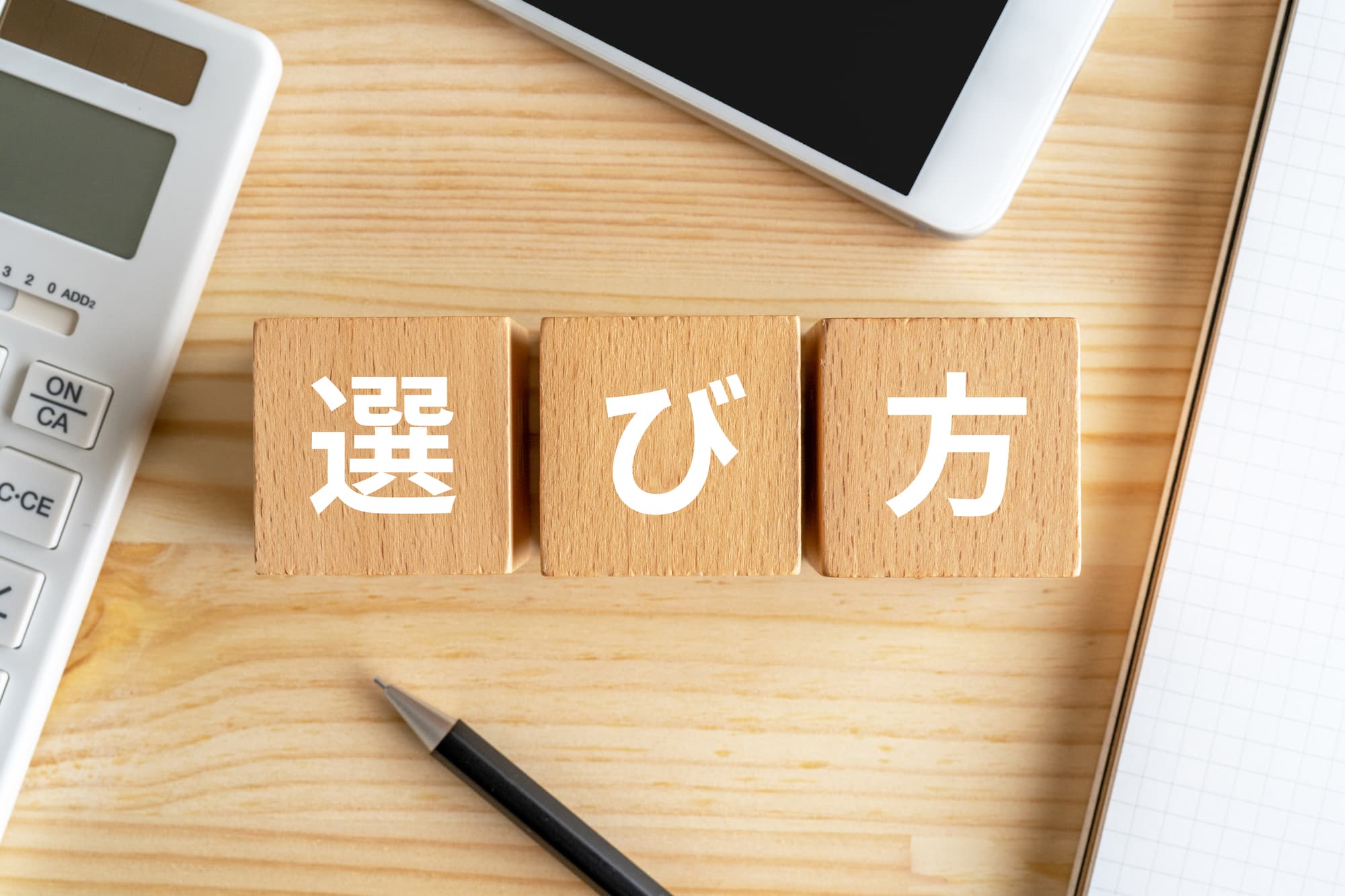 「選び方」と書かれた木のブロックとペン、電卓、スマートフォン、ノート