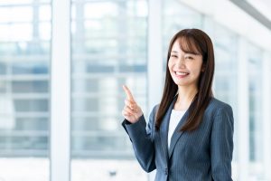 人さし指を上に向けて笑顔を浮かべるスーツ姿の女性