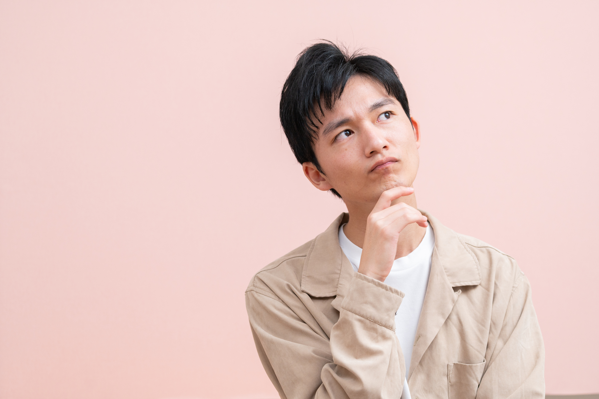 手を顎に当て斜め上を見て考えている様子の男性、ピンクの背景