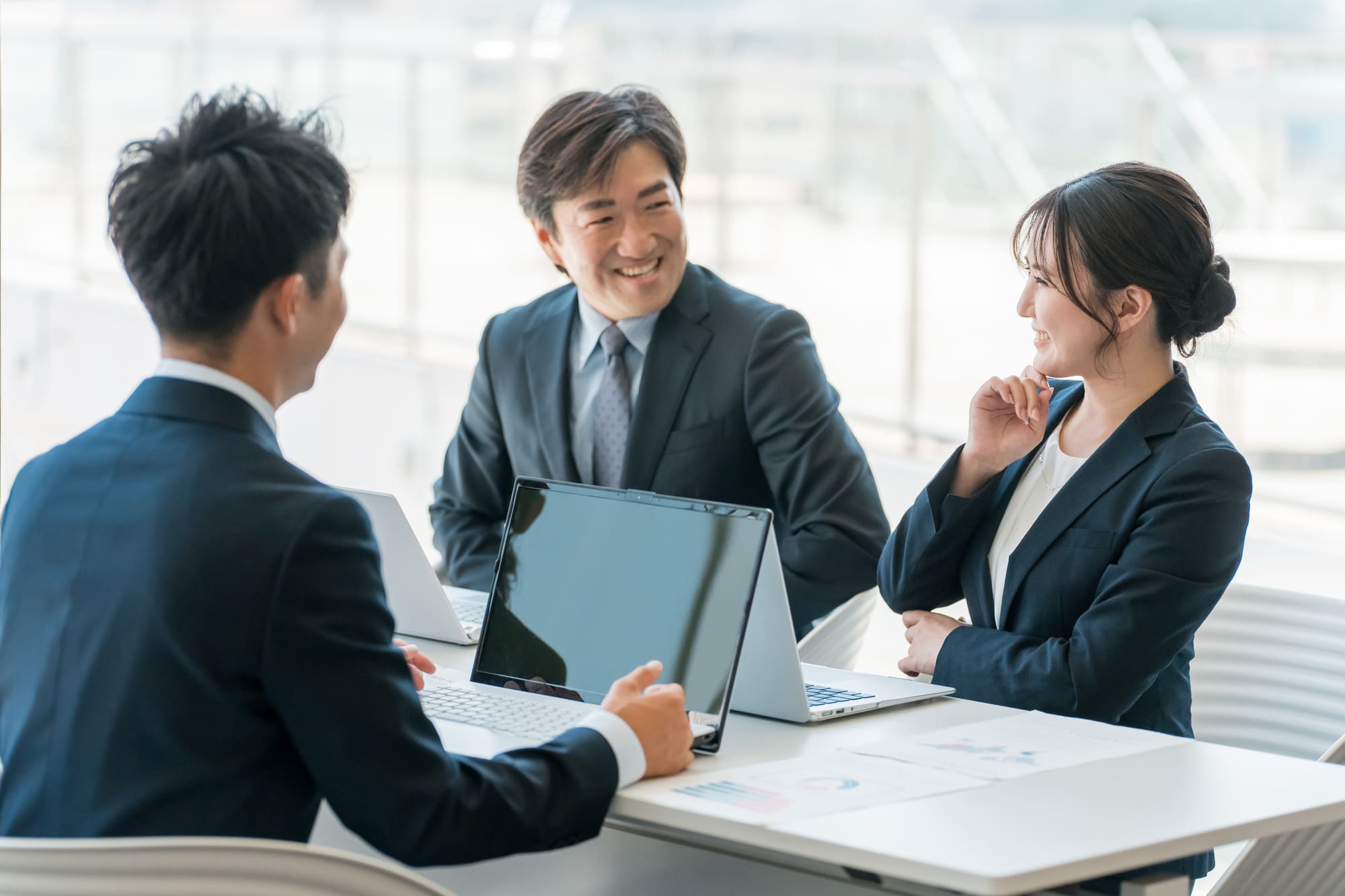 オフィスの机にノートパソコンを並べて、笑顔で会議を行う3人のビジネスパーソン