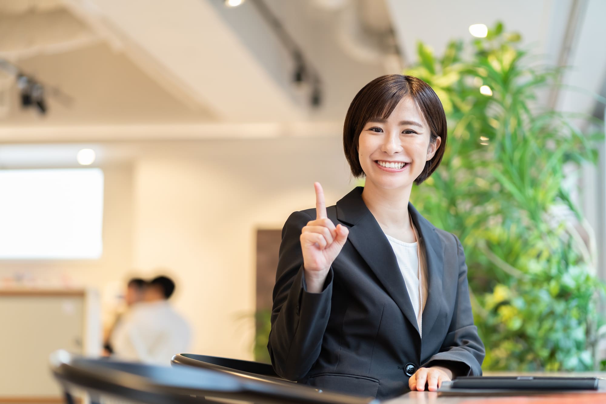人さし指を上に向け、笑顔を浮かべるスーツ姿の女性